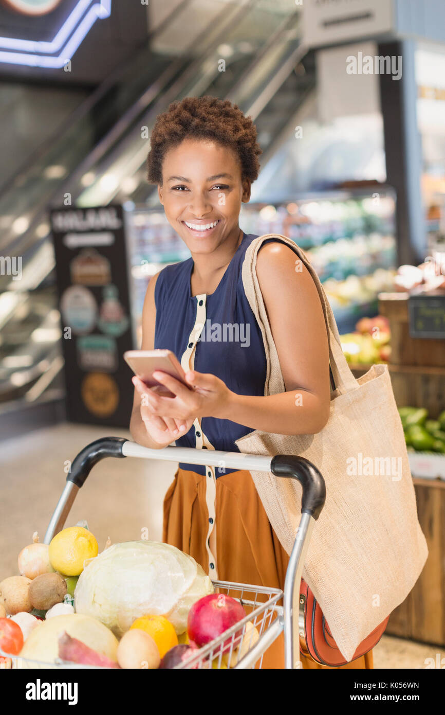 Portrait smiling young woman using cell phone, grocery shopping in market Stock Photo