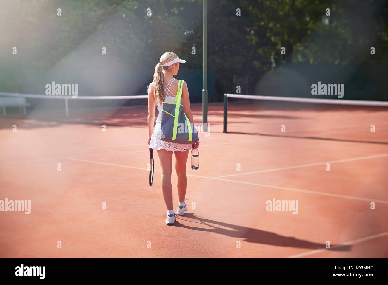 Young female tennis player walking with tennis racket, bag and water bottle on sunny clay tennis court Stock Photo