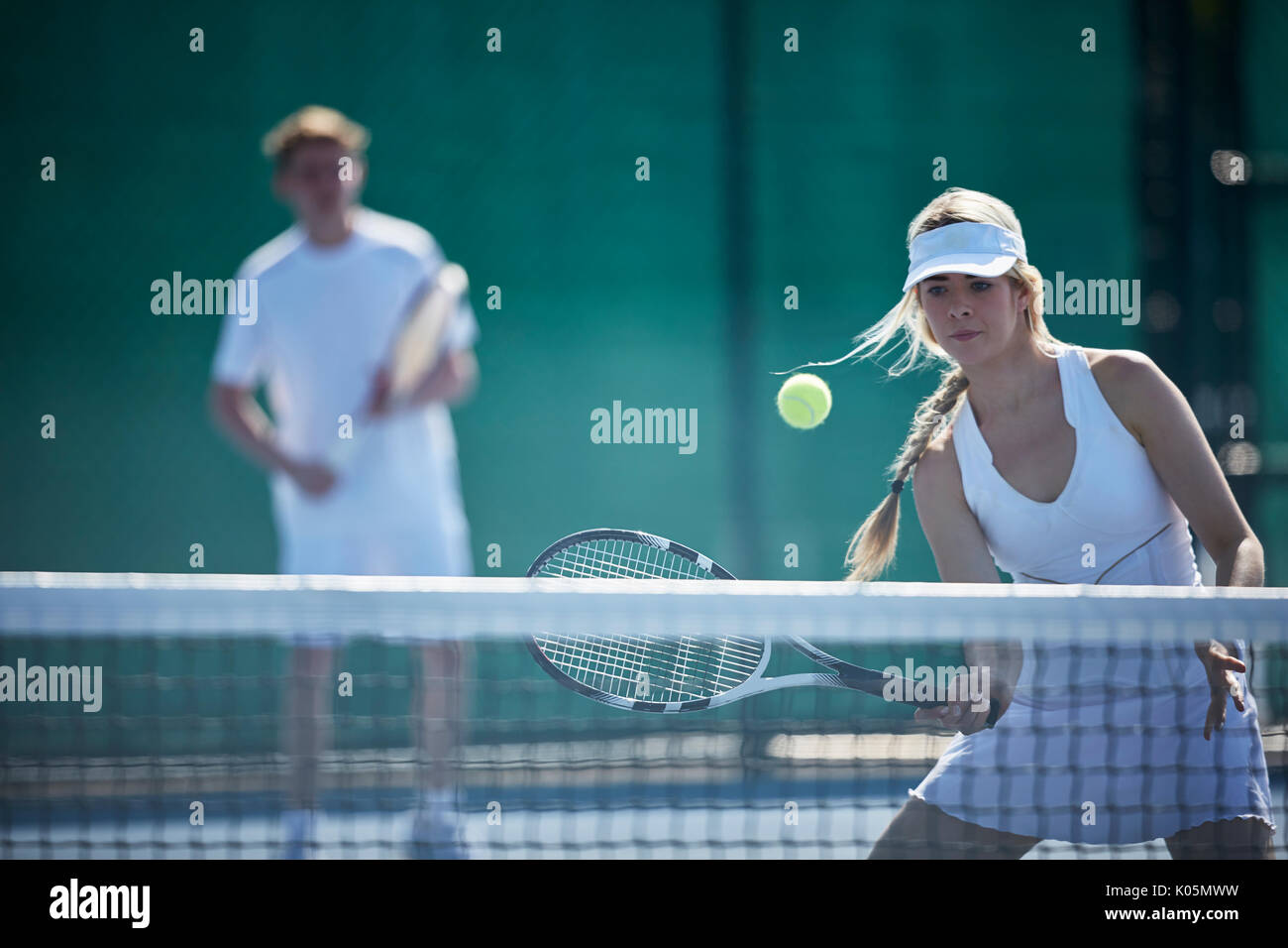 Determined young female tennis player playing tennis, hitting the ball at tennis net on sunny tennis court Stock Photo