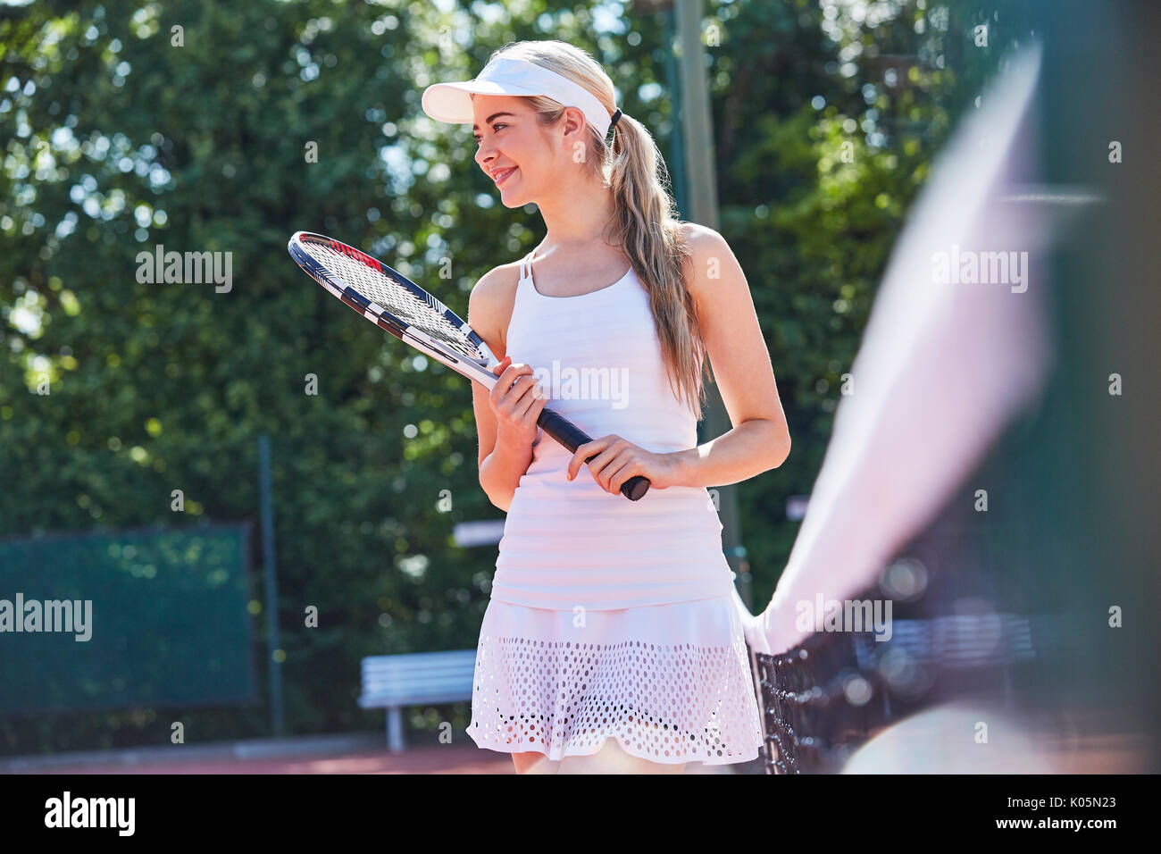 Smiling young female tennis player holding tennis racket along sunny net Stock Photo