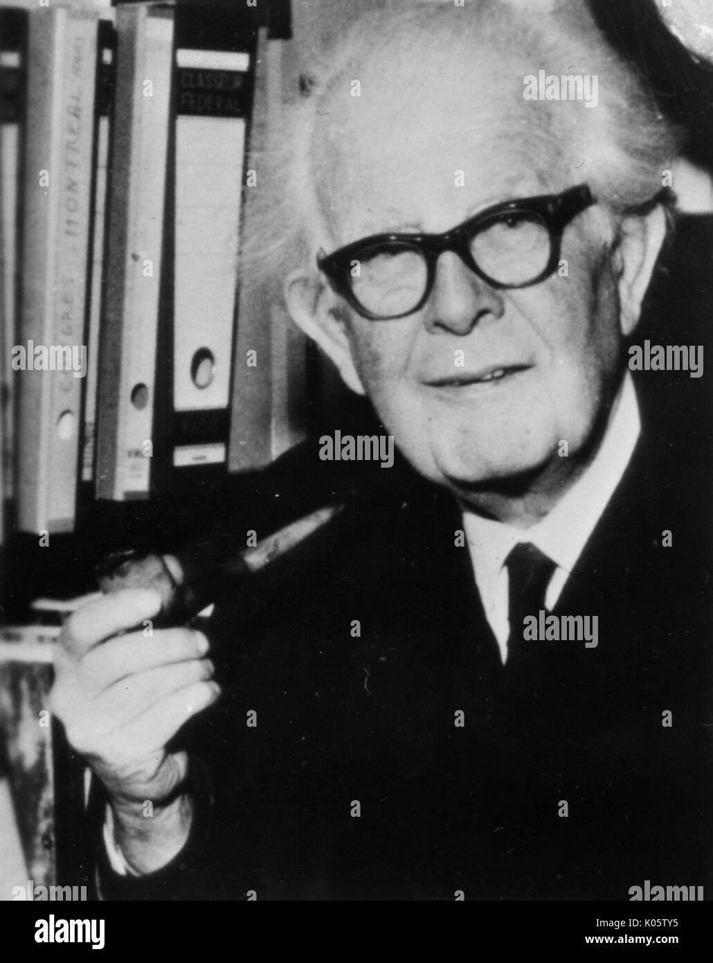 Half-length portrait of clinical psychologist Jean Piaget, wearing a dark suit and dark glasses, standing in front of a bookshelf, smoking a pipe, with a serious facial expression, 1976. Stock Photo