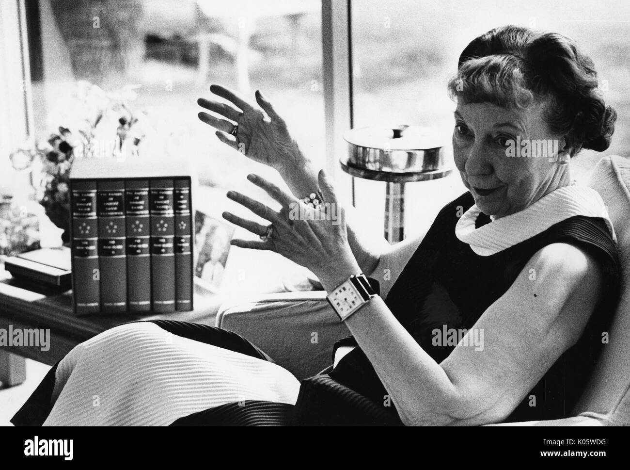 Three-quarter length portrait of Mamie Eisenhower, former First Lady of the United States and wife of former United States President Dwight Eisenhower, seated in a chair by a window, wearing a dark and white dress, gesturing both hands in the air, with a smiling facial expression, 1973. Stock Photo