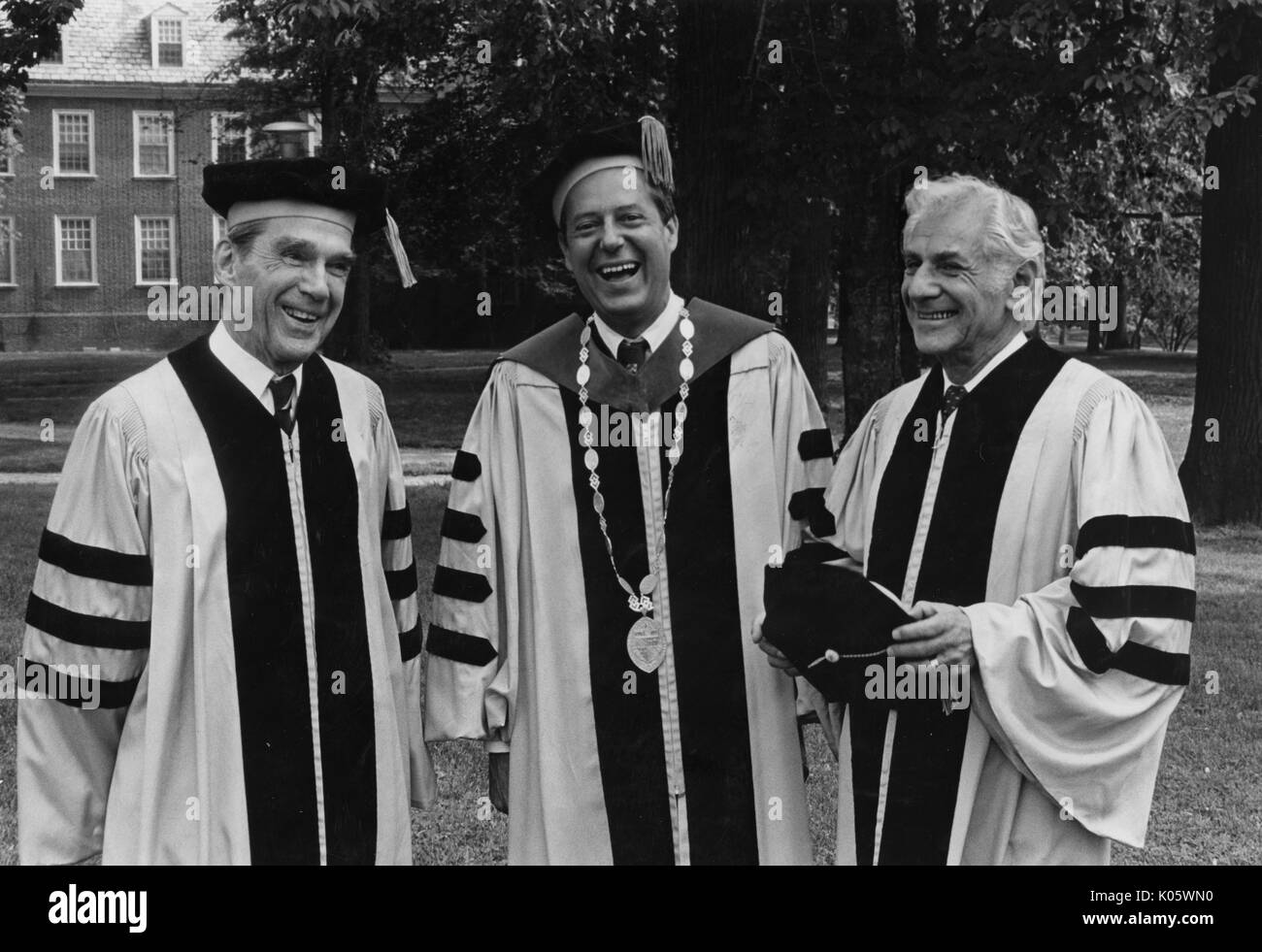 Half-body portrait of three men at Johns Hopkins commencement in 1980, all wearing cap and gown attire, standing on a campus quad, with smiling facial expressions, from left to right, Daniel K Ludwig, President Steven Muller, and musician Leonard Bernstein, May 30, 1980. Stock Photo