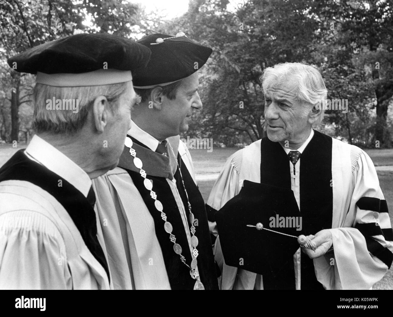 Half-body portrait of three men at 1980 Johns Hopkins commencement, all in cap and gown attire, from left to right: Daniel K Ludwig, President Steven Muller, and Leonard Bernstein, who has removed his hat and drawn the other two's attention towards him, May 30, 1980. Stock Photo