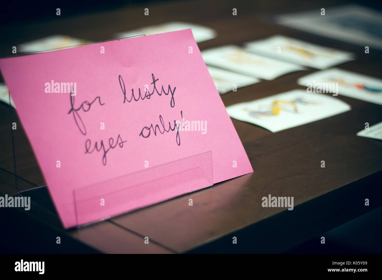 A pink sign reading 'for lusty eyes only' in black cursive writing on a wooden table with materials in soft focus behind it, 2016. Stock Photo