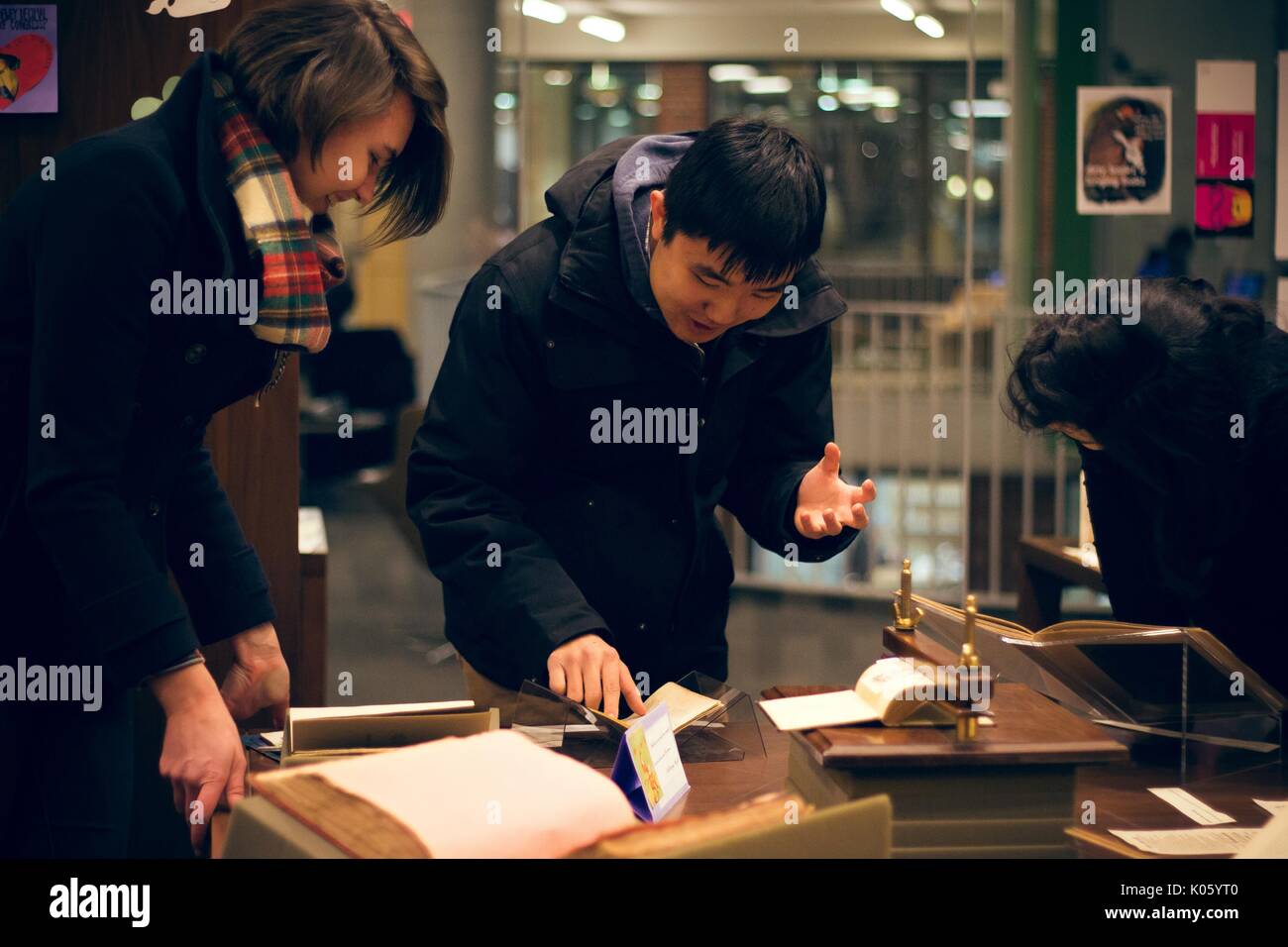 Asian male college student wearing a jacket, passionately reading aloud from a manuscript in the Special Collections department of a University library, one hand pointing to a passage in the book, the other hand gesturing, while a female student smiles and looks on, 2016. Courtesy Eric Chen. Stock Photo