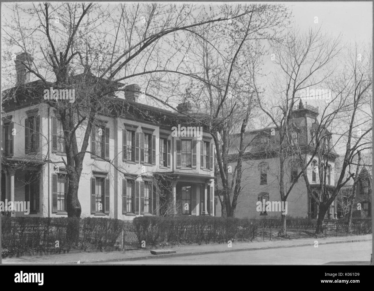 View of a street in the foreground and three homes in the background, the homes are all at least two-story and are all different designs, there are trees and bushes that line the property between the homes and the street, United States, 1910. This image is from a series documenting the construction and sale of homes in the Roland Park/Guilford neighborhood of Baltimore, a streetcar suburb and one of the first planned communities in the United States. Stock Photo