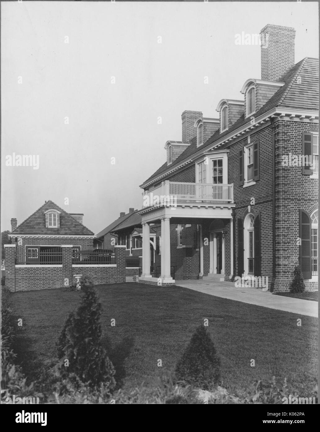 Side view of a two-story single-family home near Roland Park and Guilford, the home is made of brick and has dark-colored window shutters, the front door has a structure above it held up with two light-colored columns, above the columns is a porch to the second-story, the garage is a separate structure, United States, 1910. This image is from a series documenting the construction and sale of homes in the Roland Park/Guilford neighborhood of Baltimore, a streetcar suburb and one of the first planned communities in the United States. Stock Photo