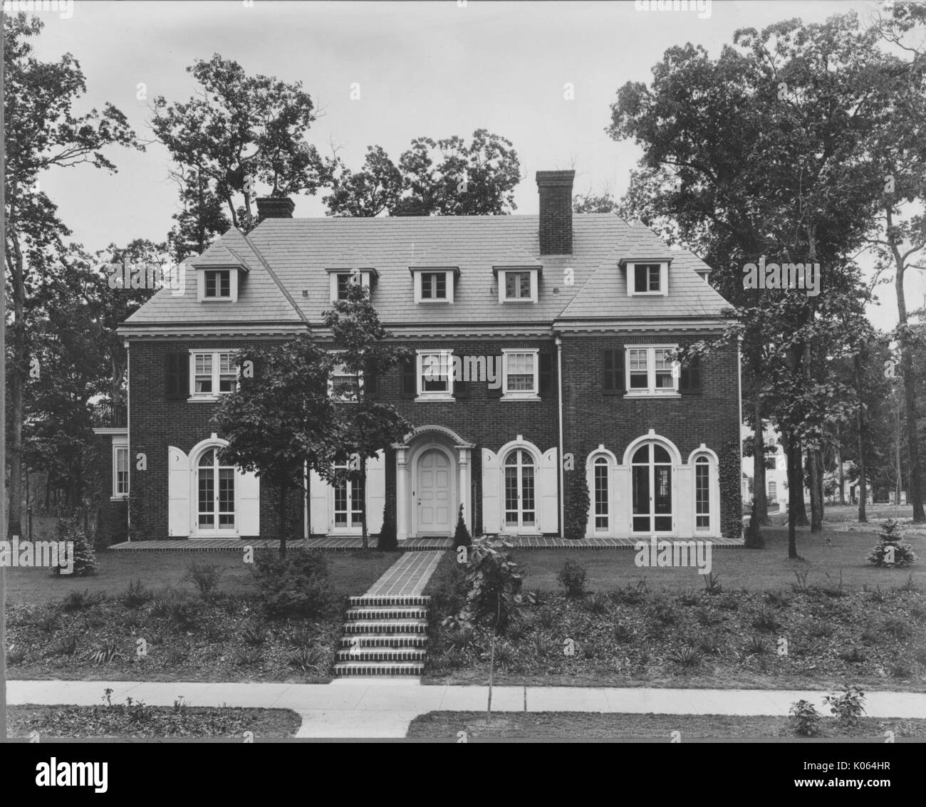 Facade view of home next to quiet street and sidewalk, brick steps and sidewalk leading up to front step of house, many windows with columns on both sides of door, two chimneys, appears to be three stories tall, many trees and small shrubs around and in front of home, United States, 1910. This image is from a series documenting the construction and sale of homes in the Roland Park/Guilford neighborhood of Baltimore, a streetcar suburb and one of the first planned communities in the United States. Stock Photo