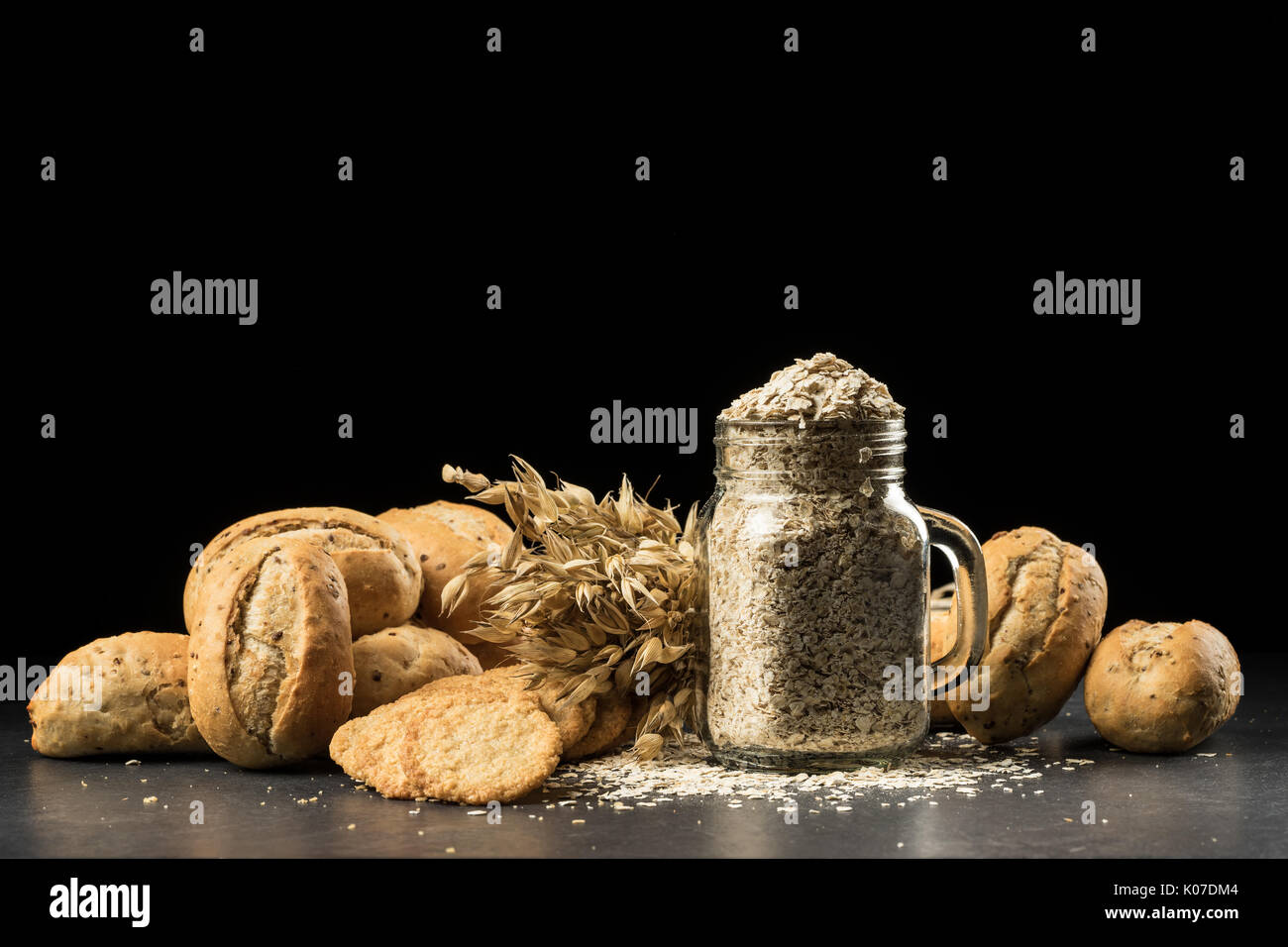 Oat bunch, baked white bread, cookies and flakes in flavouring jar, isolated on black background. Grain bouquet, golden oats spikelets on dark wooden  Stock Photo