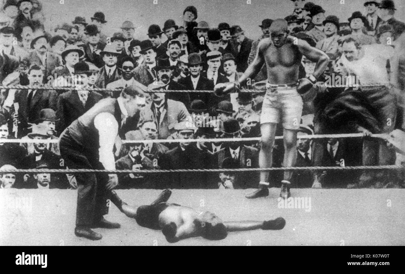 Jack Johnson (1878-1946), nicknamed the Galveston Giant, African-American world heavyweight champion boxer, 1908-1915, standing at the ropes in a boxing match with an unidentified opponent, who appears to have been knocked out.  circa 1910 Stock Photo