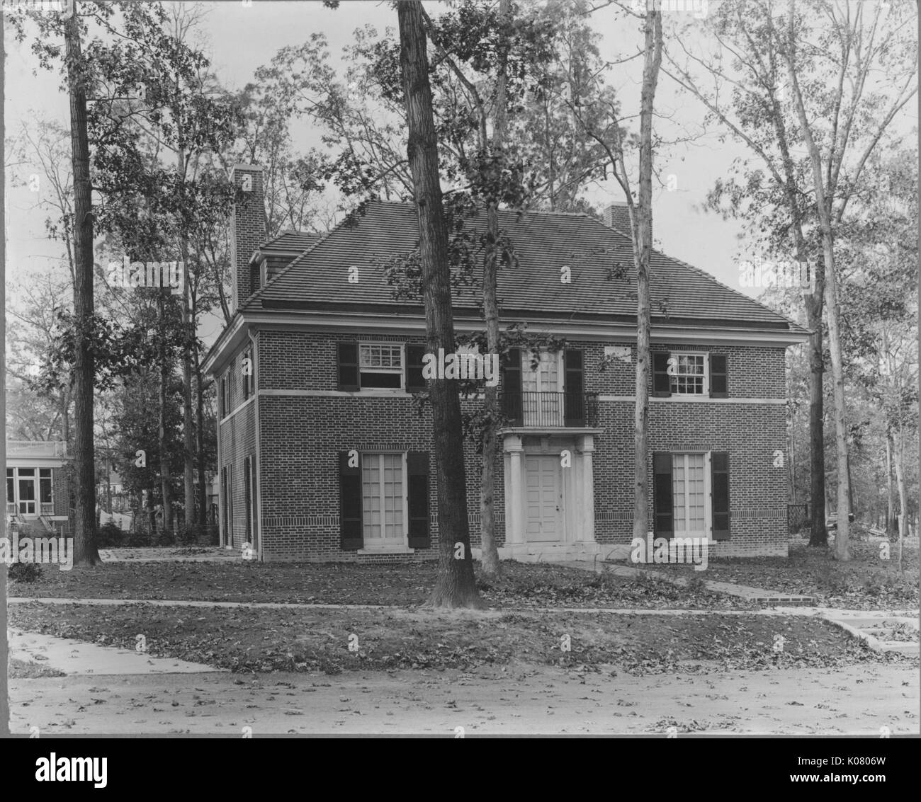 Frontal view of home next to quiet street, behind several trees, appears to be three stories tall, columns on both sides of front door, leaves scattered on the floor around home, appears to be made out of brick, United States, 1910. This image is from a series documenting the construction and sale of homes in the Roland Park/Guilford neighborhood of Baltimore, a streetcar suburb and one of the first planned communities in the United States. Stock Photo