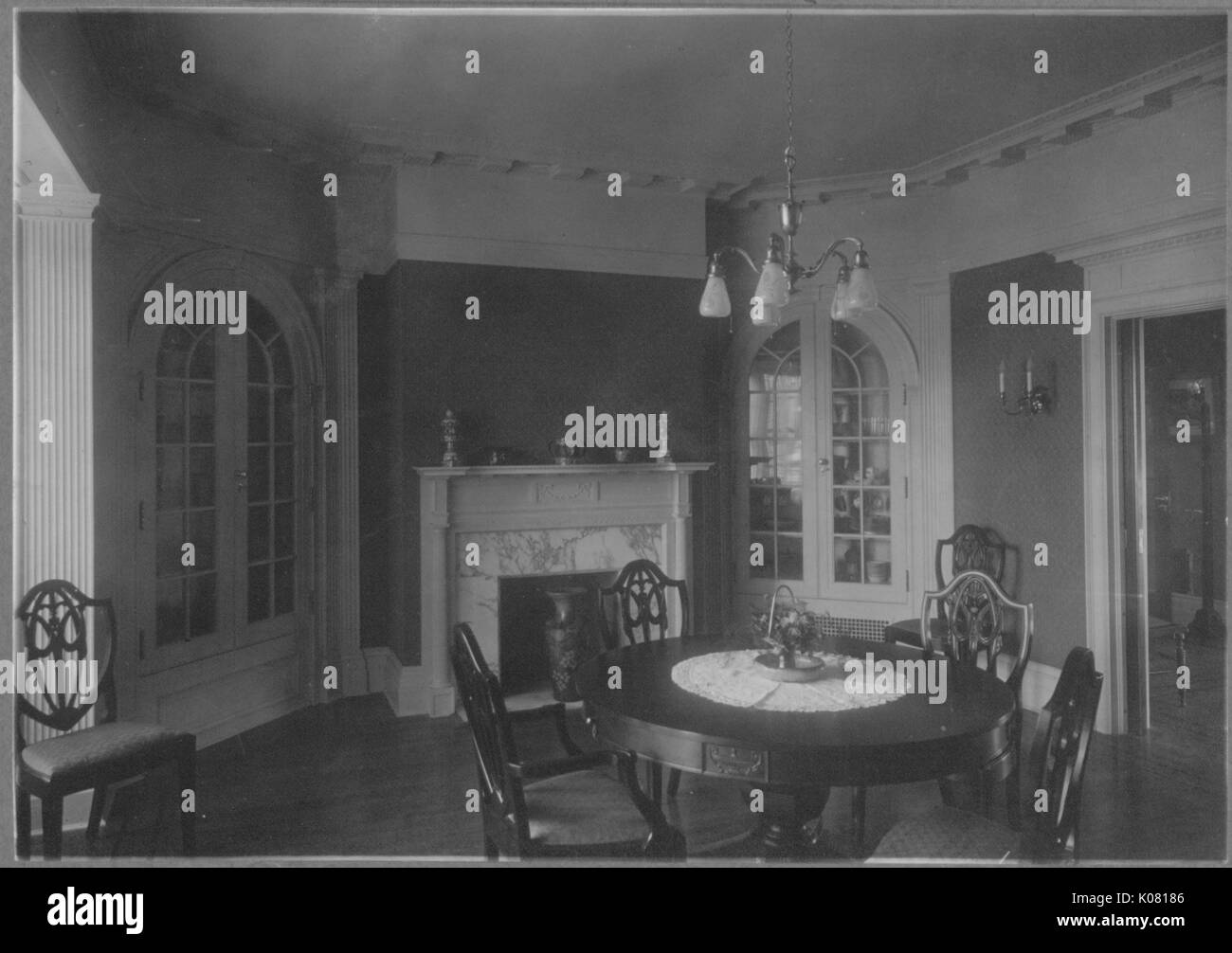 Photograph of a Roland Park dining room in Baltimore, with a round wooden table and wooden chairs around it, with an ornate white fireplace, with a wooden floor, and two large white glass cabinets and a small chandelier hanging above the table, United States, 1920. This image is from a series documenting the construction and sale of homes in the Roland Park/Guilford neighborhood of Baltimore, a streetcar suburb and one of the first planned communities in the United States. Stock Photo