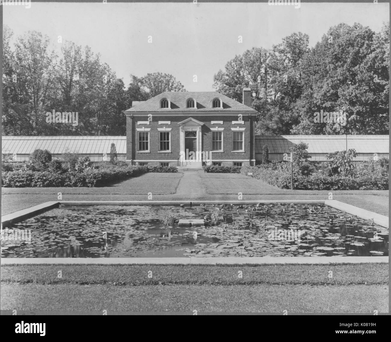 Photograph of the exterior of a brick home featuring standard and dormer windows, a small staircase leading to an entryway, and chimneys, situated in an elaborately landscaped lawn with a reflecting pool, dense trees behind the home, in a quiet residential area of Baltimore, Maryland, 1910. This image is from a series documenting the construction and sale of homes in the Roland Park/Guilford neighborhood of Baltimore, a streetcar suburb and one of the first planned communities in the United States. Stock Photo