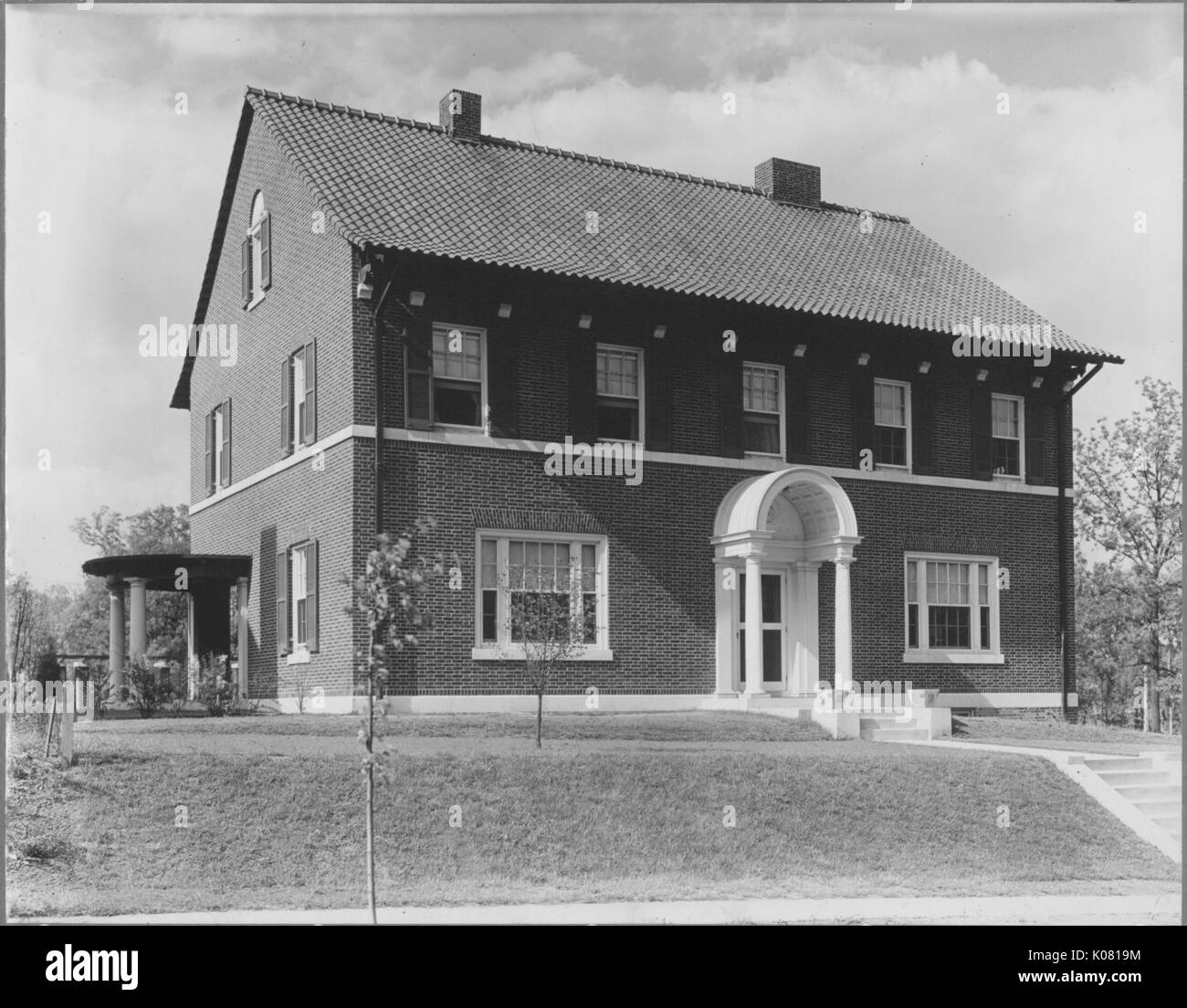 Photograph of the exterior of a brick home in Baltimore, Maryland featuring windows with and without shutters, chimneys, and two small sets of stairs leading to a covered entryway, situated in a sparsely landscapes lawn on a residential street with a sidewalk in Baltimore, Maryland, 1910. This image is from a series documenting the construction and sale of homes in the Roland Park/Guilford neighborhood of Baltimore, a streetcar suburb and one of the first planned communities in the United States. Stock Photo