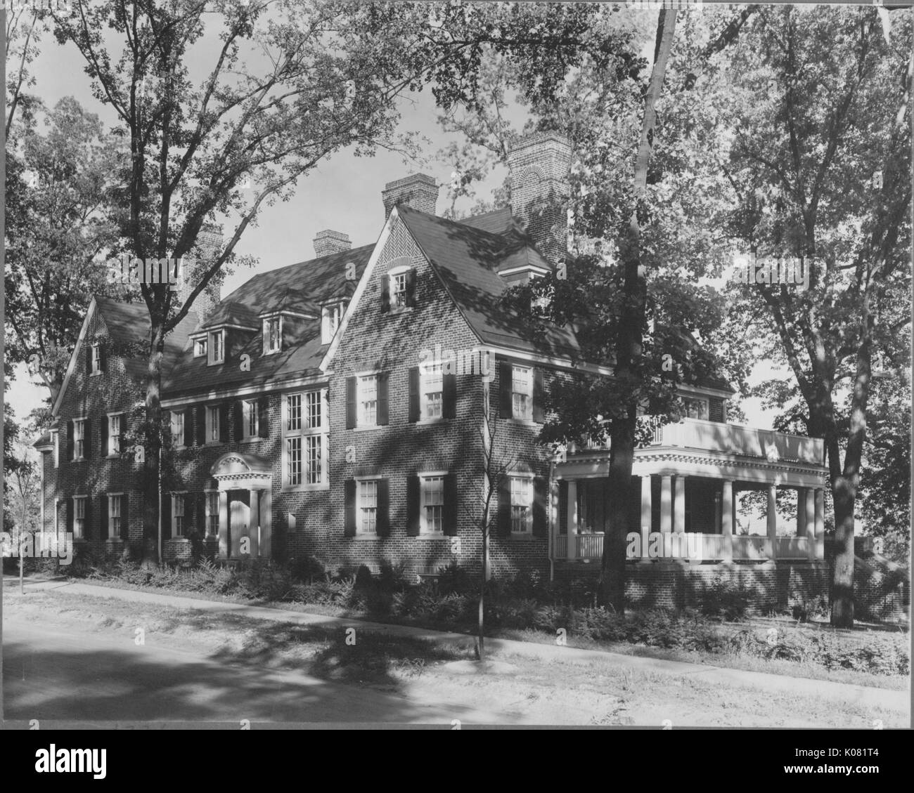 Photograph of a large Roland Park home made of brick, with two large balconies, a two-story home many windows, no front yard, the porch has two Doric columns, and the home is shaded by many leafy trees, United States, 1950. This image is from a series documenting the construction and sale of homes in the Roland Park/Guilford neighborhood of Baltimore, a streetcar suburb and one of the first planned communities in the United States. Stock Photo