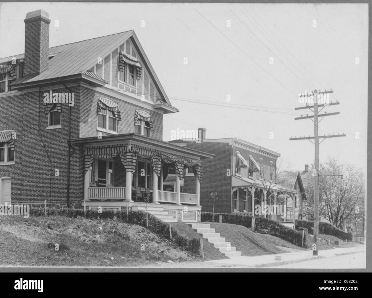 Photograph of a street in Baltimore with Roland Park homes, risen above the street, both brick homes with large front porches in between low hedges, United States, 1920. This image is from a series documenting the construction and sale of homes in the Roland Park/Guilford neighborhood of Baltimore, a streetcar suburb and one of the first planned communities in the United States. Stock Photo