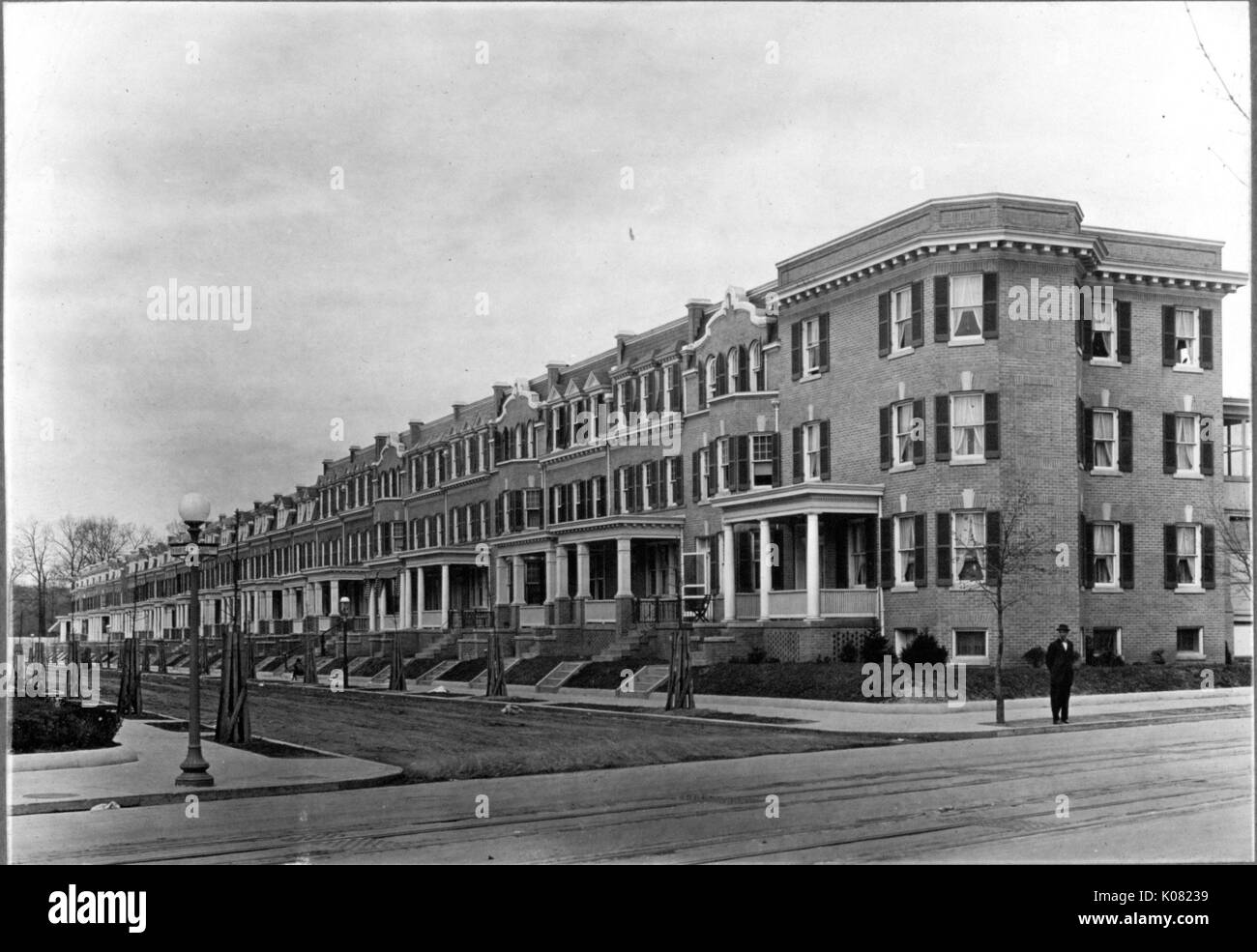 A street with a sidewalk on which a man stands by a line of row houses, each with a small set of stairs leading to a front porch and entryway, shuttered windows, and a chimney, Baltimore, Maryland, 1910. This image is from a series documenting the construction and sale of homes in the Roland Park/Guilford neighborhood of Baltimore, a streetcar suburb and one of the first planned communities in the United States. Stock Photo