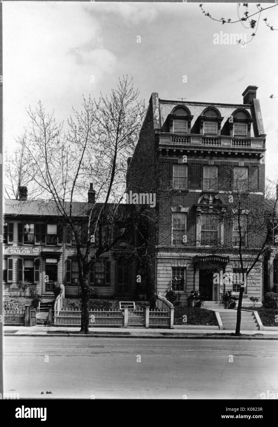 Two buildings sit on a street wit a tree-lined sidewalk in Baltimore, Maryland, one (left) with the sign for a tailor, shrubbery, a short fence, shuttered windows, and chimneys; the other (right) brick, with a small staircase leading to the entryway, shrubbery, a chimney, top-story dormer windows, and ornate stone accents, Baltimore, Maryland, 1910. This image is from a series documenting the construction and sale of homes in the Roland Park/Guilford neighborhood of Baltimore, a streetcar suburb and one of the first planned communities in the United States. Stock Photo