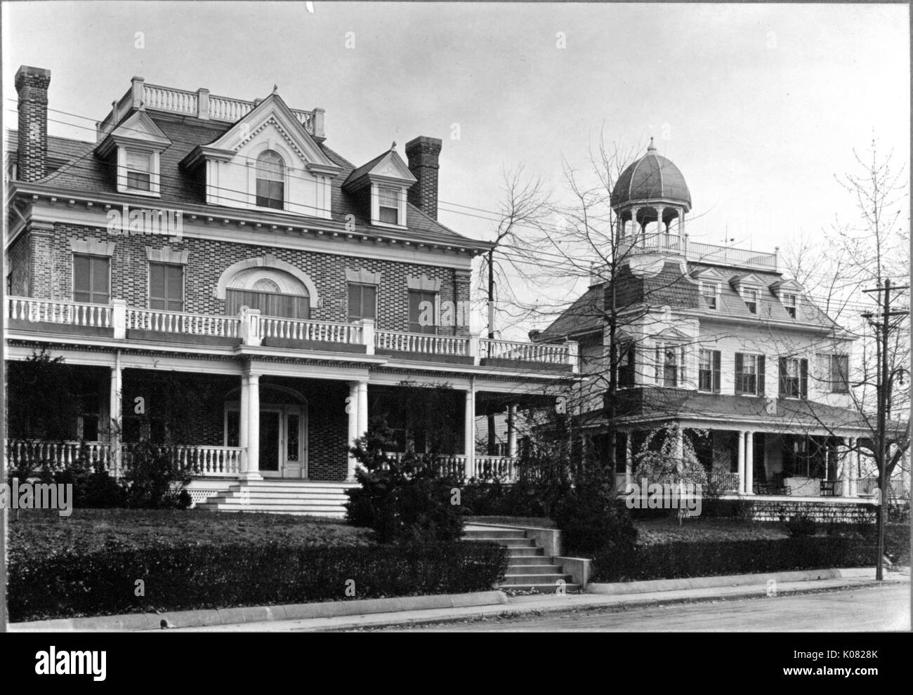 Two homes sit on a residential street in Baltimore, Maryland; both with hedge shrubbery, one (left) brick with first-story and second-story wrap-around porches, two small sets of steps leading to the front entryway, top-story dormer windows, two chimneys, and a rooftop porch; the other (right) with a front porch, shuttered windows, top-story dormer windows, and a roof porch and gazebo, Baltimore, Maryland, 1910. This image is from a series documenting the construction and sale of homes in the Roland Park/Guilford neighborhood of Baltimore, a streetcar suburb and one of the first planned commun Stock Photo