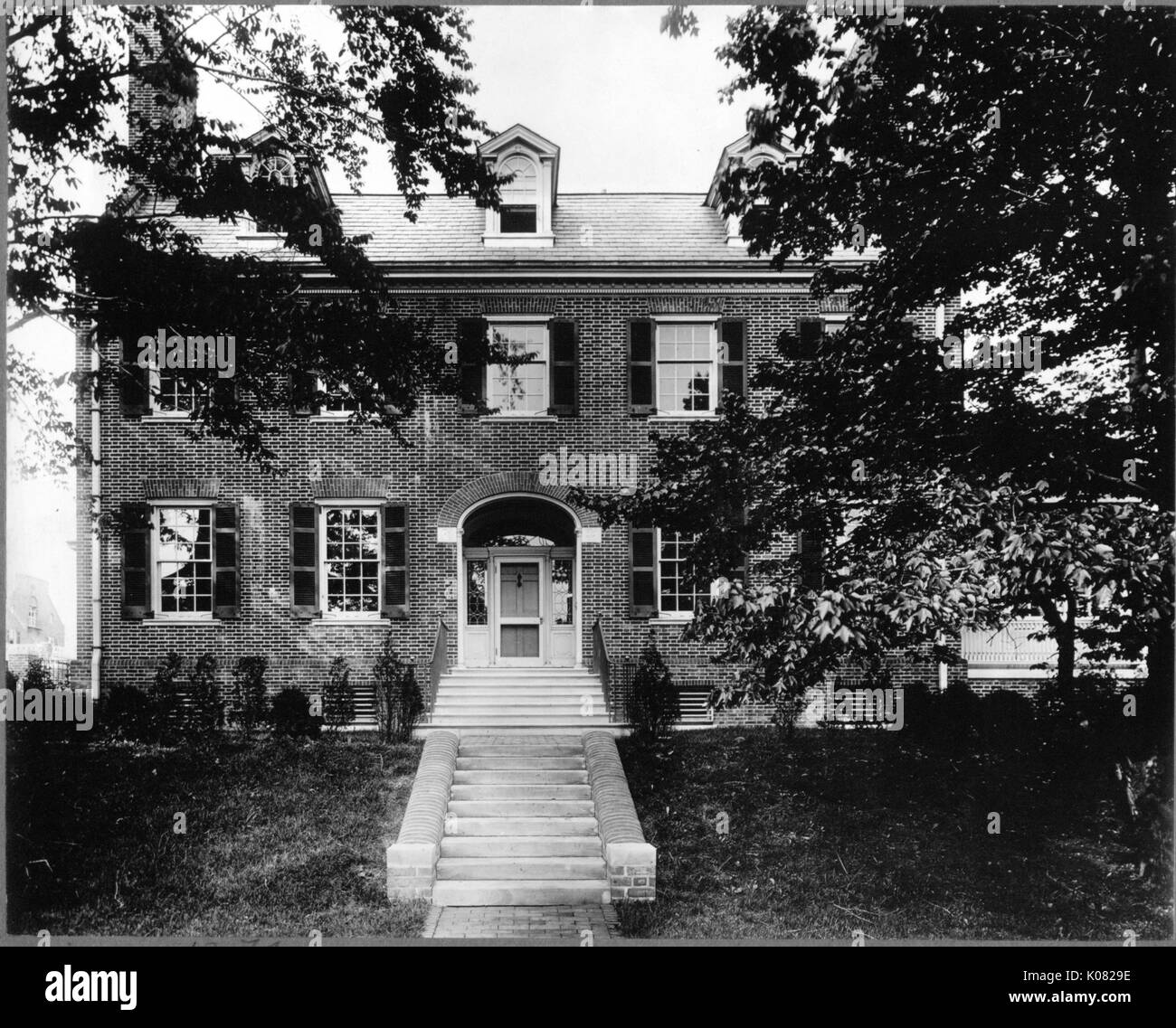 A large, brick home with a two small sets of stairs leading to an arched entryway, windows with shutters, dormer windows, an end chimney, and an obscured side porch (right) sits in a yard with trees and shrubbery in Baltimore, Maryland, 1910. This image is from a series documenting the construction and sale of homes in the Roland Park/Guilford neighborhood of Baltimore, a streetcar suburb and one of the first planned communities in the United States. Stock Photo