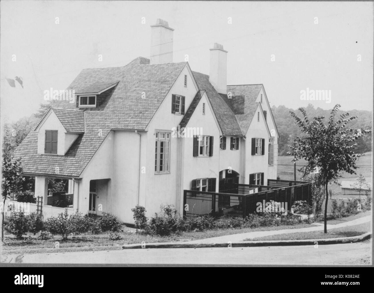 Angled side view of home on quiet street, part of home covered by fence, front porch visible from angle, house surrounded by vegetation such as bushes and trees, two chimneys coming out of top of house, windows with dark panels, house next to concrete sidewalk and street, Baltimore, Maryland, 1910. This image is from a series documenting the construction and sale of homes in the Roland Park/Guilford neighborhood of Baltimore, a streetcar suburb and one of the first planned communities in the United States. Stock Photo