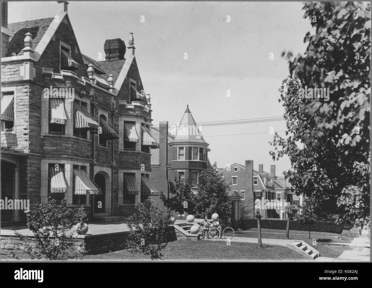 Angled view of homes on quiet street, cobblestone home with striped coverings for shade on windows, triangular shapes in top of homes, chimneys on top of homes, house has front porch with bicycles and steps, bushes and trees surrounding home, concrete sidewalk on side of homes, Baltimore, Maryland, 1910. This image is from a series documenting the construction and sale of homes in the Roland Park/Guilford neighborhood of Baltimore, a streetcar suburb and one of the first planned communities in the United States. Stock Photo