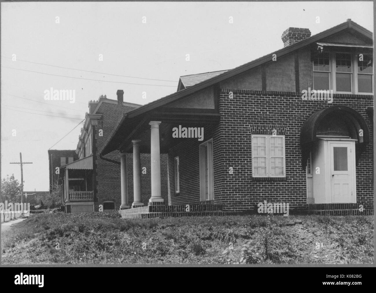 Side view of brick homes on quiet street surrounded by vegetation, House in front has front porch and side door and columns, telephone poles, wires and trees around house, Baltimore, Maryland, 1910. This image is from a series documenting the construction and sale of homes in the Roland Park/Guilford neighborhood of Baltimore, a streetcar suburb and one of the first planned communities in the United States. Stock Photo