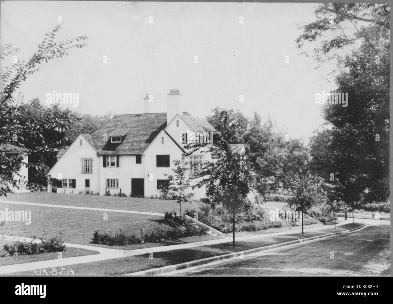 View of side of light-colored house in showing several windows and door with dark panels, chimney on house, fields of grass and concrete sidewalks surrounding house, much vegetation and trees lining house and sidewalks, street next to house, Baltimore, Maryland, 1910. This image is from a series documenting the construction and sale of homes in the Roland Park/Guilford neighborhood of Baltimore, a streetcar suburb and one of the first planned communities in the United States. Stock Photo