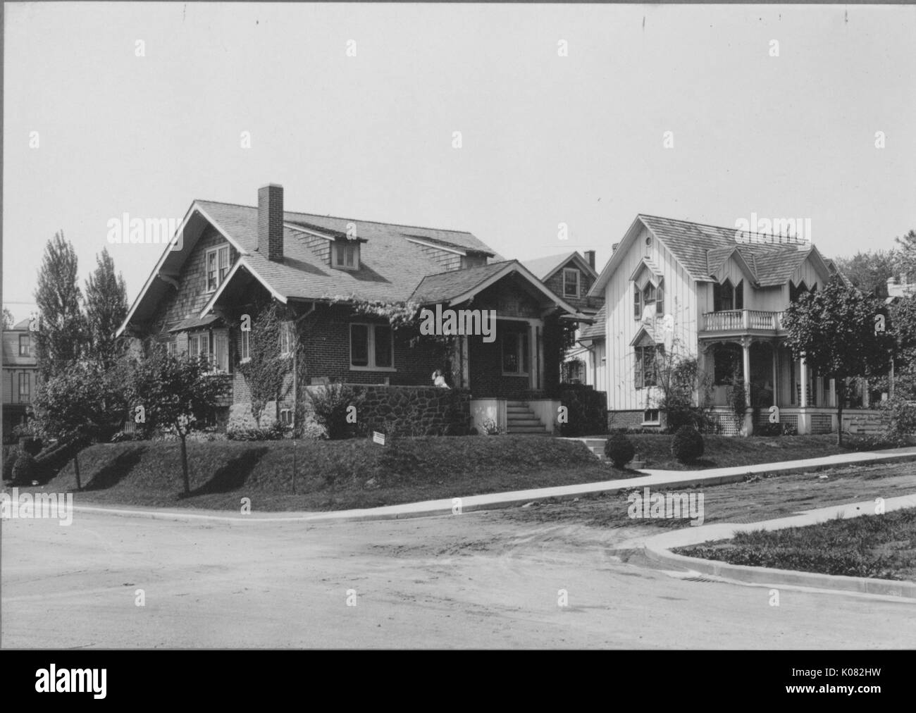 Angled side view of two homes on quiet street, one house made of dark-colored stone and brick with chimney and plants surrounding house and vines alongside wall, another house with light-colored panels, porch and columns, street and sidewalks running alongside homes, trees lining sidewalks, houses on slight hill, Baltimore, Maryland, 1910. This image is from a series documenting the construction and sale of homes in the Roland Park/Guilford neighborhood of Baltimore, a streetcar suburb and one of the first planned communities in the United States. Stock Photo