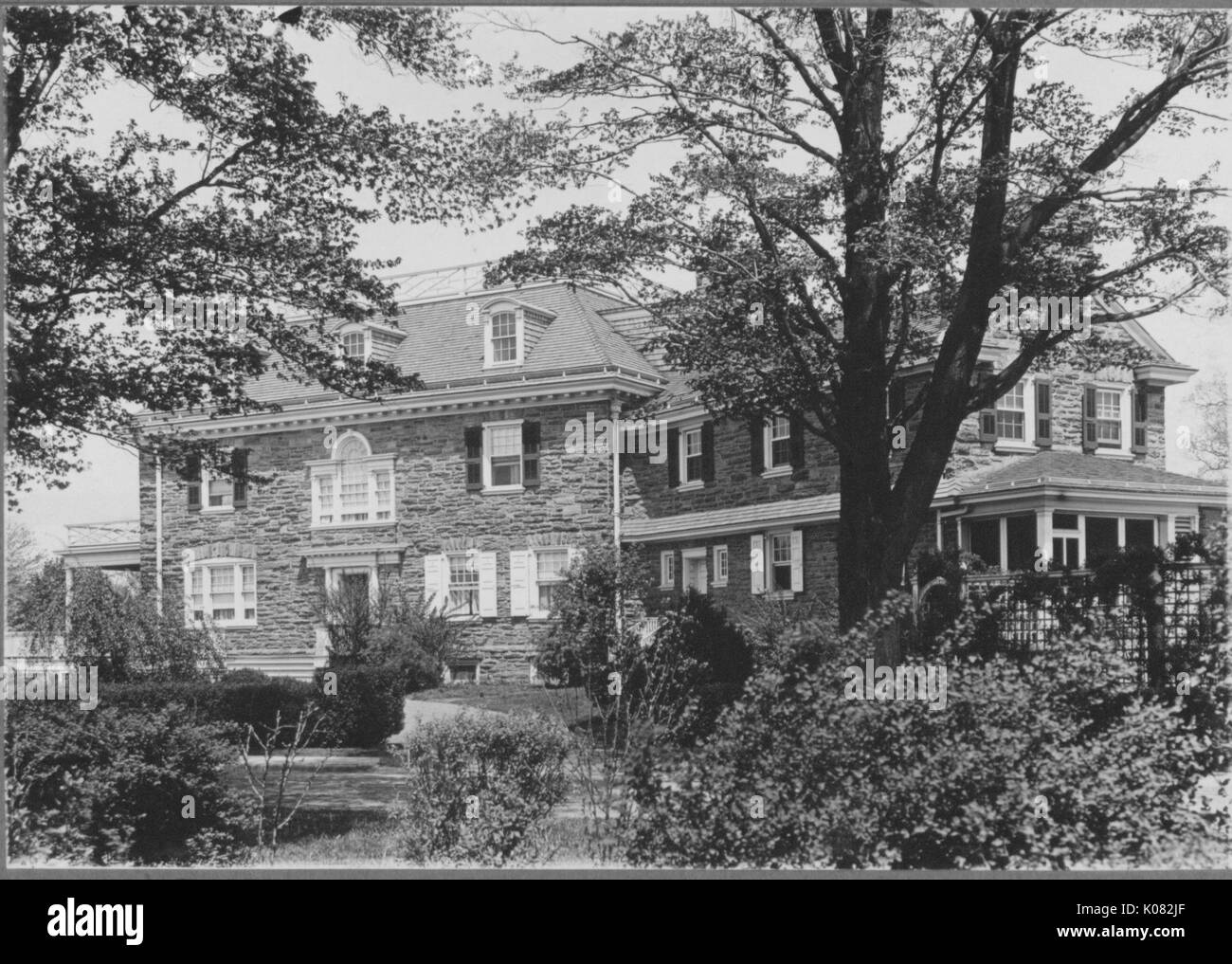 Angled view of large home with cobblestone walls and many windows, house has porch, view of home covered by wide variety of vegetation from bushes to trees, Baltimore, Maryland, 1910. This image is from a series documenting the construction and sale of homes in the Roland Park/Guilford neighborhood of Baltimore, a streetcar suburb and one of the first planned communities in the United States. Stock Photo