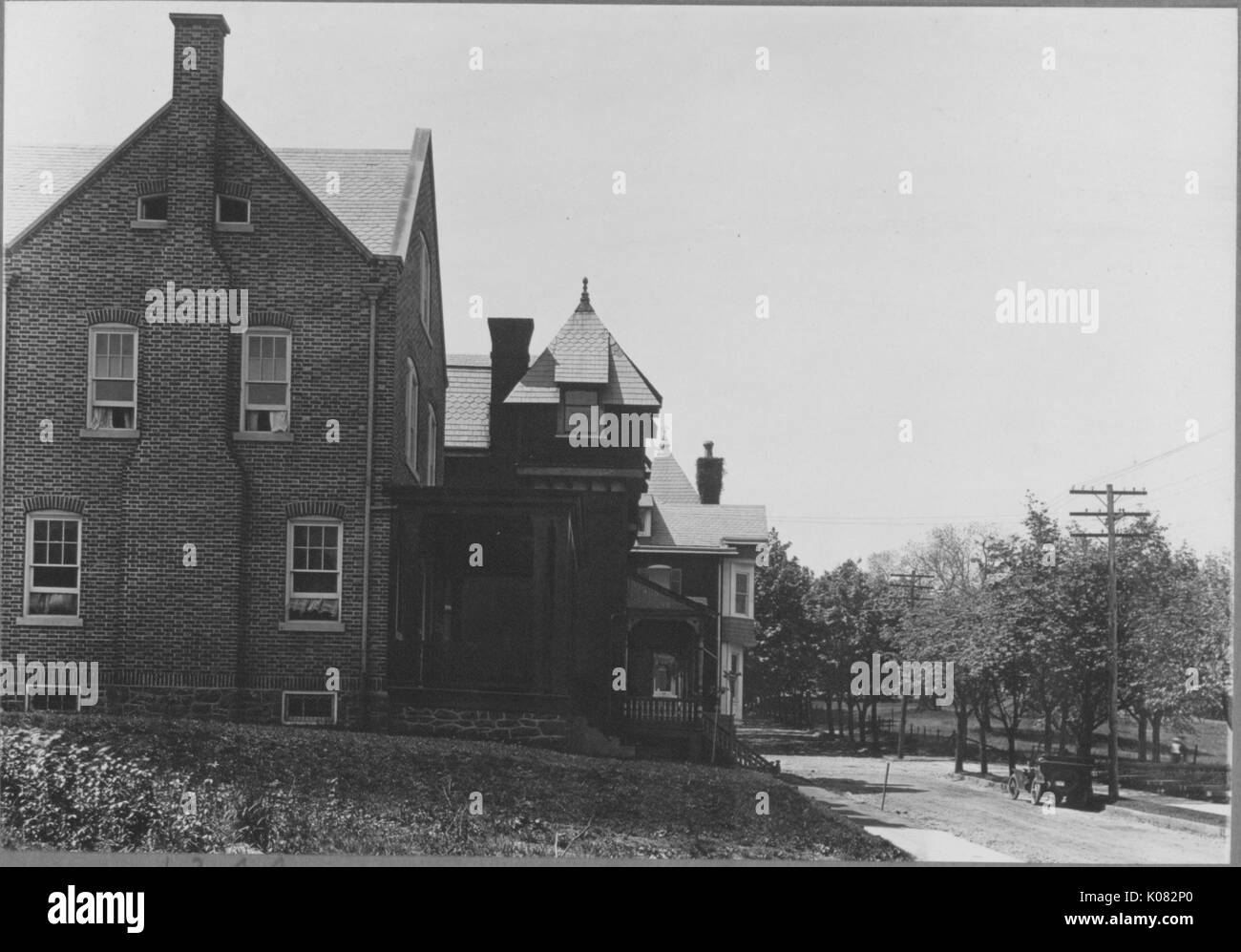 Side view of brick house on quiet street, house has four large windows on side, chimney and front porch, front porch overlooks trees, vacant spot with vegetation on side of house, Baltimore, Maryland, 1910. This image is from a series documenting the construction and sale of homes in the Roland Park/Guilford neighborhood of Baltimore, a streetcar suburb and one of the first planned communities in the United States. Stock Photo