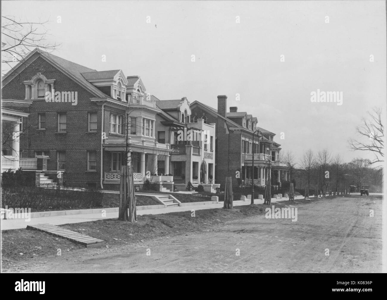 Diagonal shot of row of houses with two stories, steps leading up to complex front porch structures, chimneys depicted, on an unpaved road; Roland Park/Guilford, 1910. This image is from a series documenting the construction and sale of homes in the Roland Park/Guilford neighborhood of Baltimore, a streetcar suburb and one of the first planned communities in the United States. Stock Photo