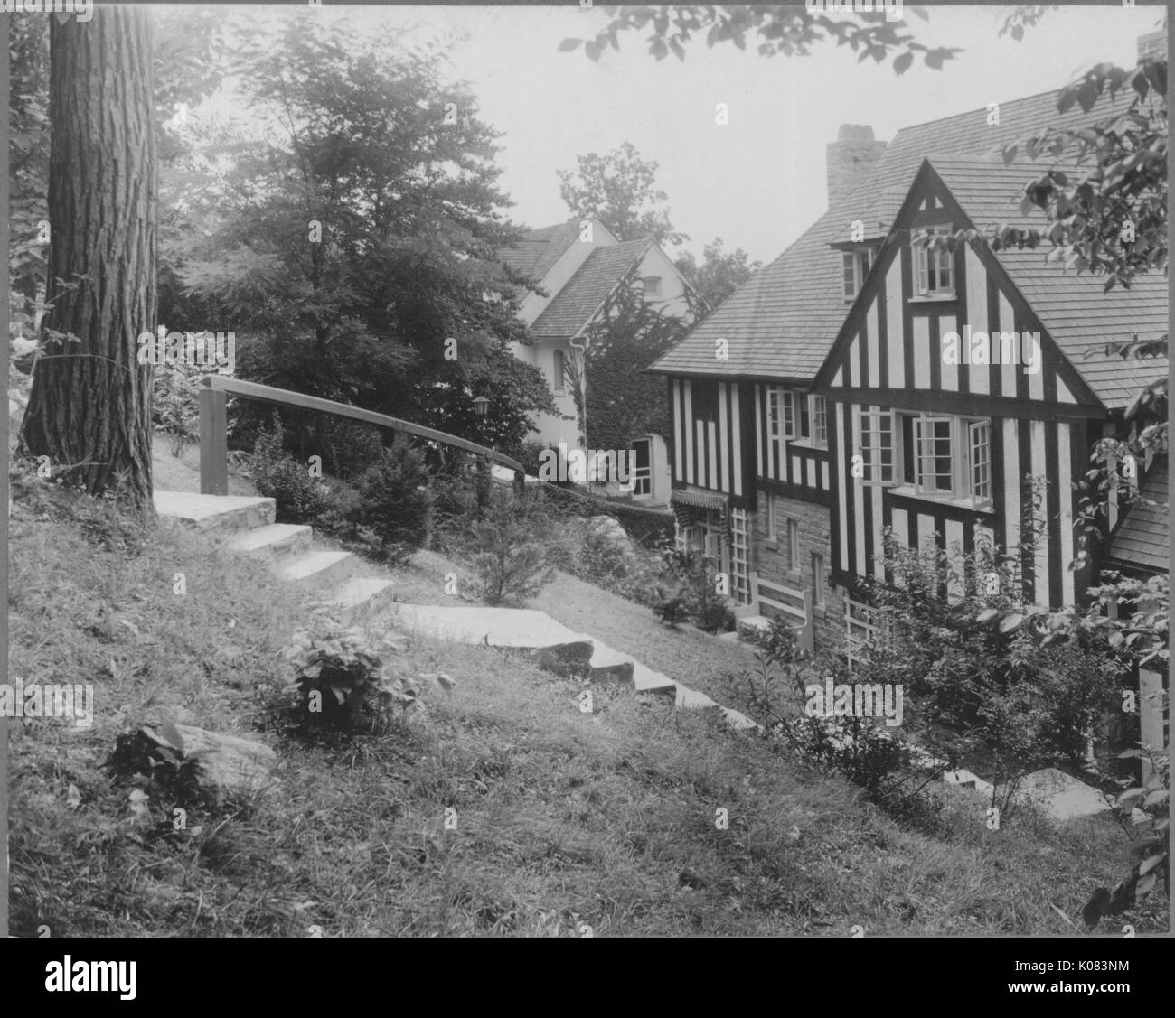 Partial view of a half-timbered and stone home with a chimney, a dormer window, standard windows, and a long set of steps with a railing leading to its entrance, set among trees and shrubbery, next to another home, partially-visible, in Baltimore, Maryland, 1910. This image is from a series documenting the construction and sale of homes in the Roland Park/Guilford neighborhood of Baltimore, a streetcar suburb and one of the first planned communities in the United States. Stock Photo