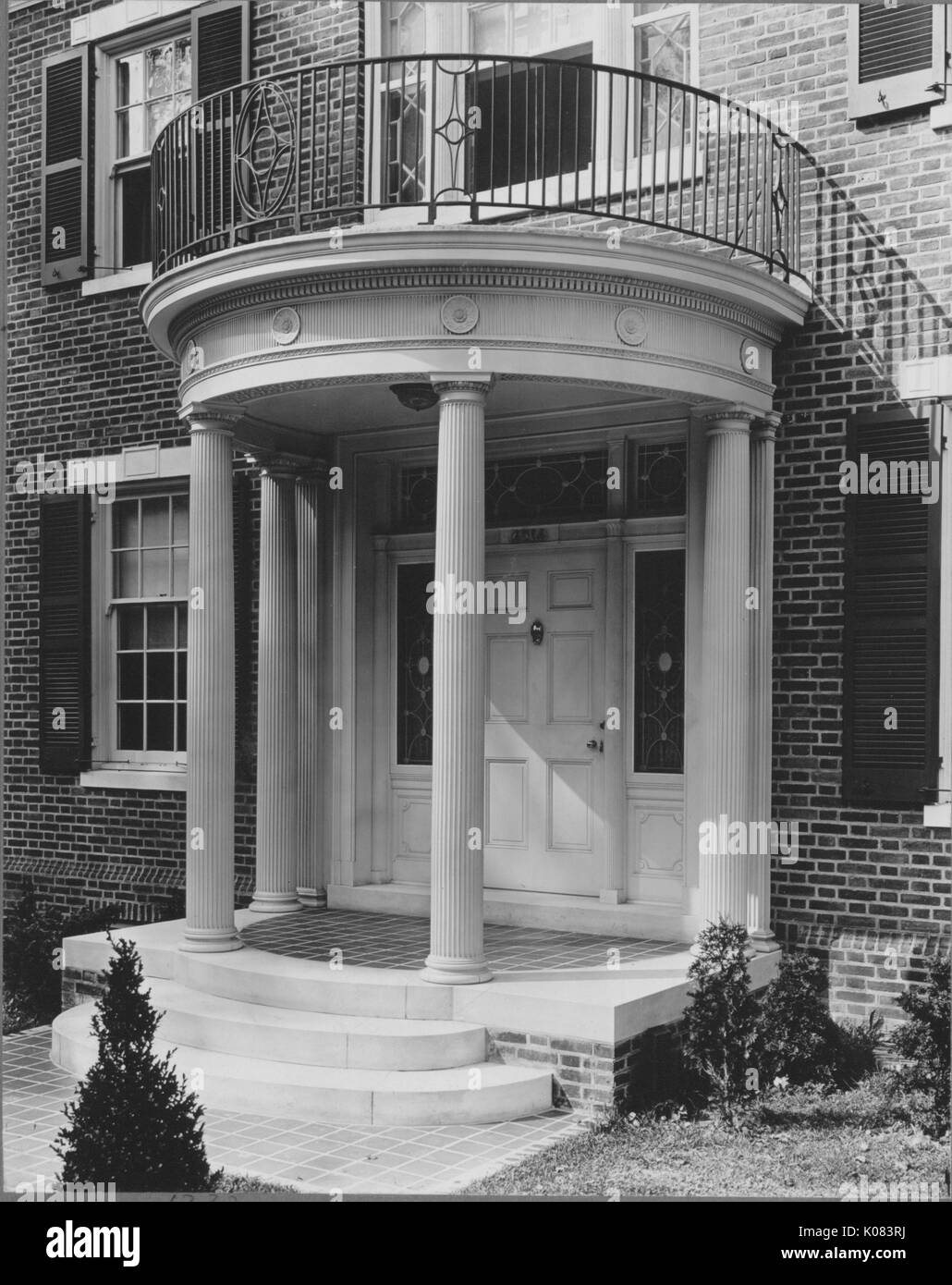 Partial view of a brick home that features windows with shutters, a second-story front balcony held up by classical columns that form the entryway to the home, a set of three steps that lead to the front door, and sparse landscaping including shrubbery, in Baltimore, Maryland, 1910. This image is from a series documenting the construction and sale of homes in the Roland Park/Guilford neighborhood of Baltimore, a streetcar suburb and one of the first planned communities in the United States. Stock Photo