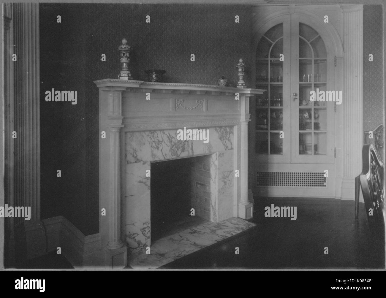 Interior of Baltimore home, featuring base molding, patterned wallpaper, a marble fireplace with a mantelpiece on which sit decorative goods, a built-in corner cabinet with various tableware on display, and a partial view of two ornate wooden chairs, Baltimore, Maryland, 1910. This image is from a series documenting the construction and sale of homes in the Roland Park/Guilford neighborhood of Baltimore, a streetcar suburb and one of the first planned communities in the United States. Stock Photo