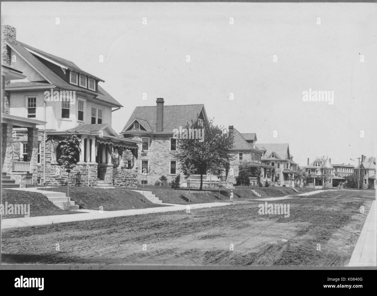 Several houses lining a quiet residential street in Baltimore, Maryland, with a sidewalk running parallel to the street, each home made at least partially of stone, each featuring a front porch, dormer windows, a chimney, and a small set of stairs leading to its yard, Baltimore, Maryland, 1910. This image is from a series documenting the construction and sale of homes in the Roland Park/Guilford neighborhood of Baltimore, a streetcar suburb and one of the first planned communities in the United States. Stock Photo
