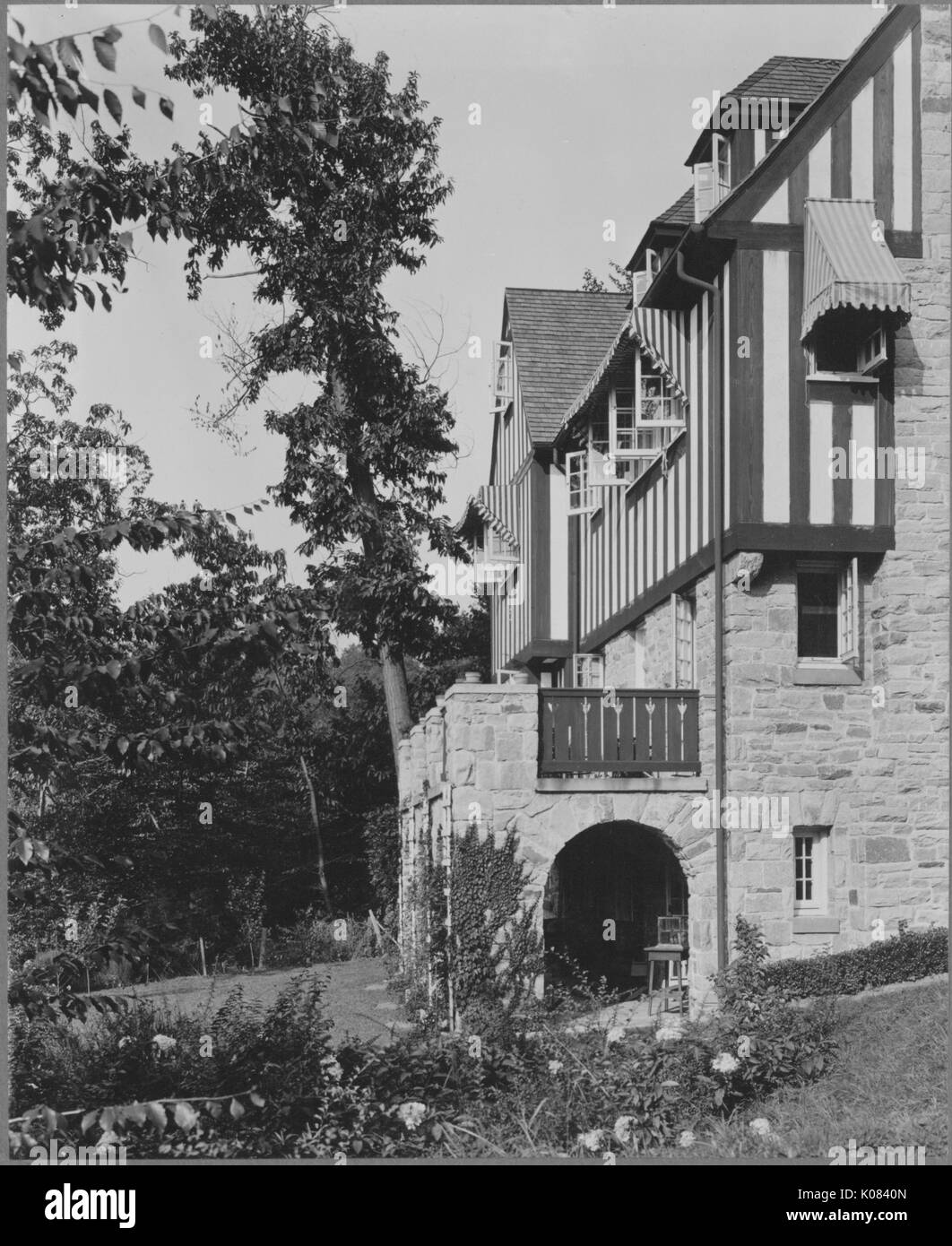 Partial view of a half-timbered and stone home taken from the side, featuring upper-story windows with striped awnings, narrow windows throughout, a second-story balcony, a first-story porch with stone arches, set in a landscaped yard with trees, shrubbery, and flowers, in Baltimore, Maryland, 1910. This image is from a series documenting the construction and sale of homes in the Roland Park/Guilford neighborhood of Baltimore, a streetcar suburb and one of the first planned communities in the United States. Stock Photo