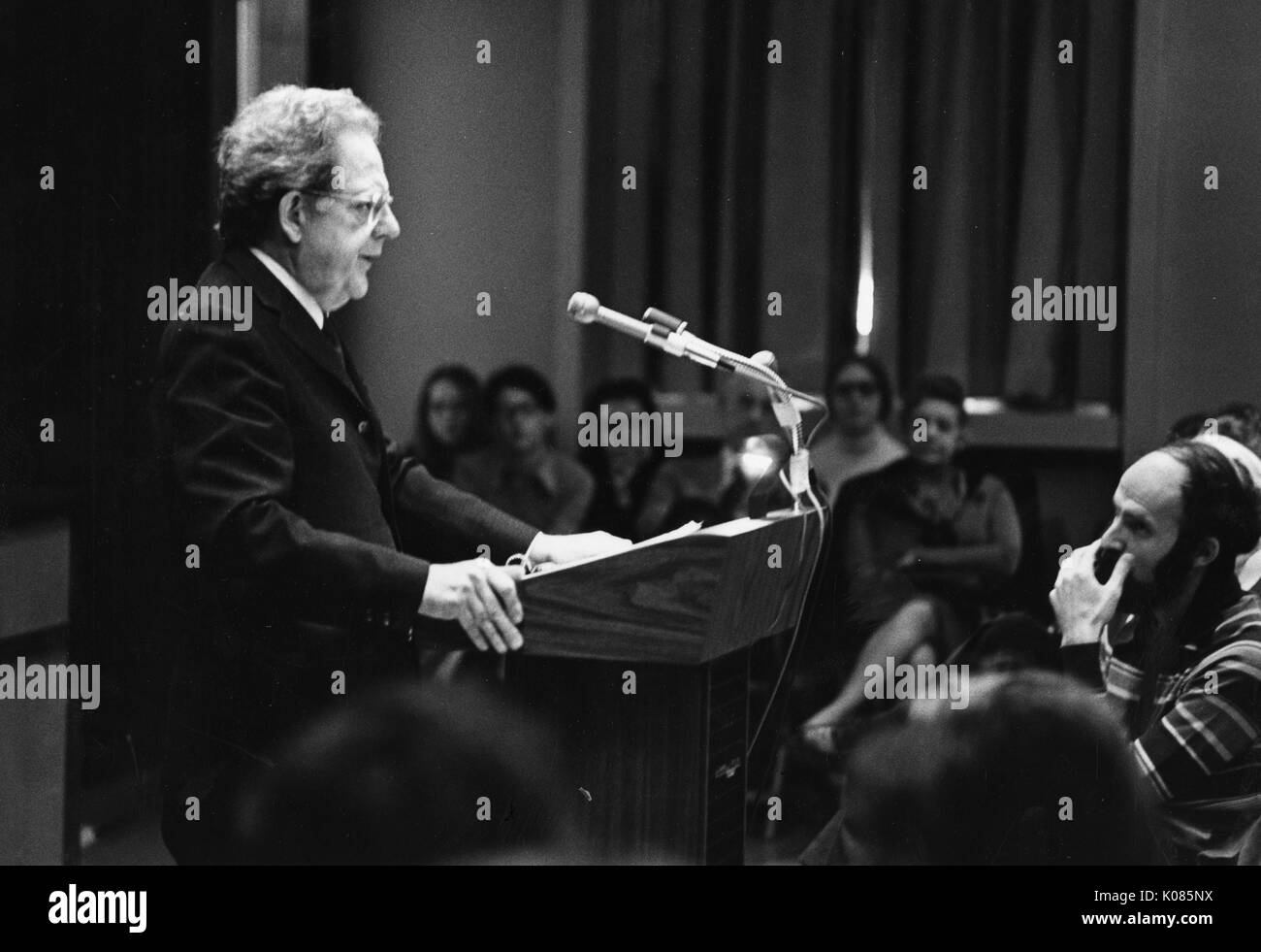 Three-quarter length portrait of author Northrup Frye, standing behind a wooden podium and making a speech to an audience, wearing a dark suit and glasses, with a serious facial expression, hands both firmly pressed on the podium, 1972. Stock Photo
