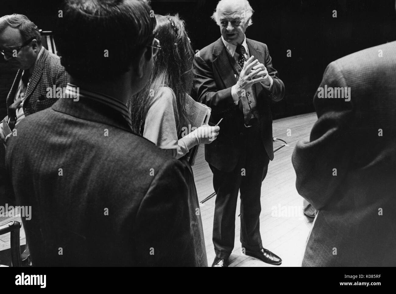 Full-length portrait of an unidentified speaker at a symposium, surrounded by people and speaking to a woman, the man wearing a dark suit and white shirt with a patterned tie, his hands clasped together in front of him, with a serious facial expression, 1976. Stock Photo