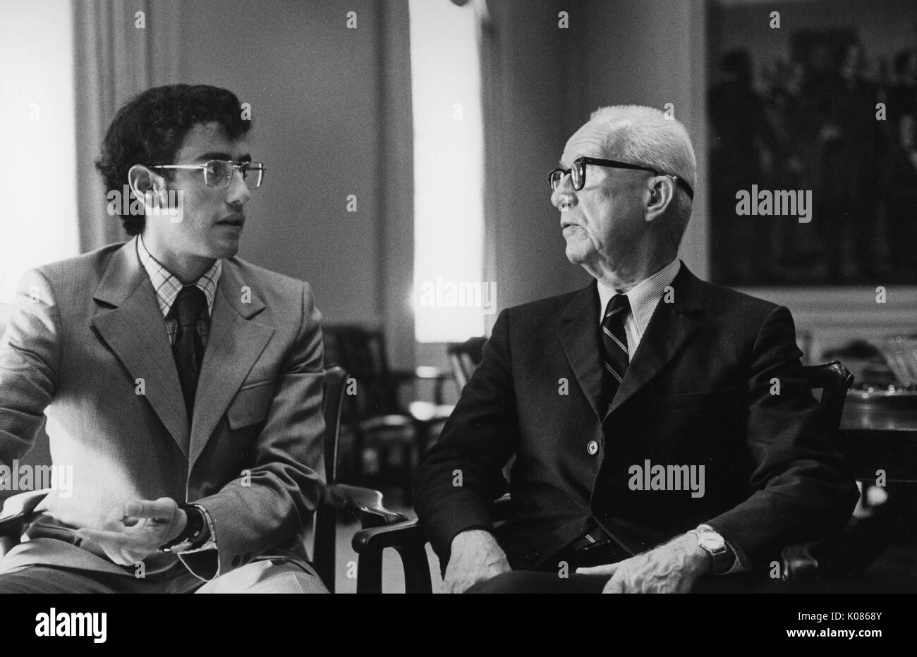 Half-length portrait of architect Buckminster Fuller speaking with another man, Fuller wearing a dark suit with a white shirt and a striped tie, wearing glasses, seated in a wooden chair and facing the other man, 1970. Stock Photo