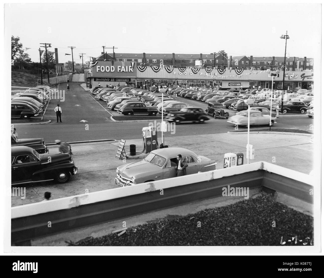 View of a gas station with pumps connected to cars, behind the gas station is a packed parking lot for the Food Fair store, behind the Food Fair store are row homes, Baltimore, Maryland, 1951. Stock Photo