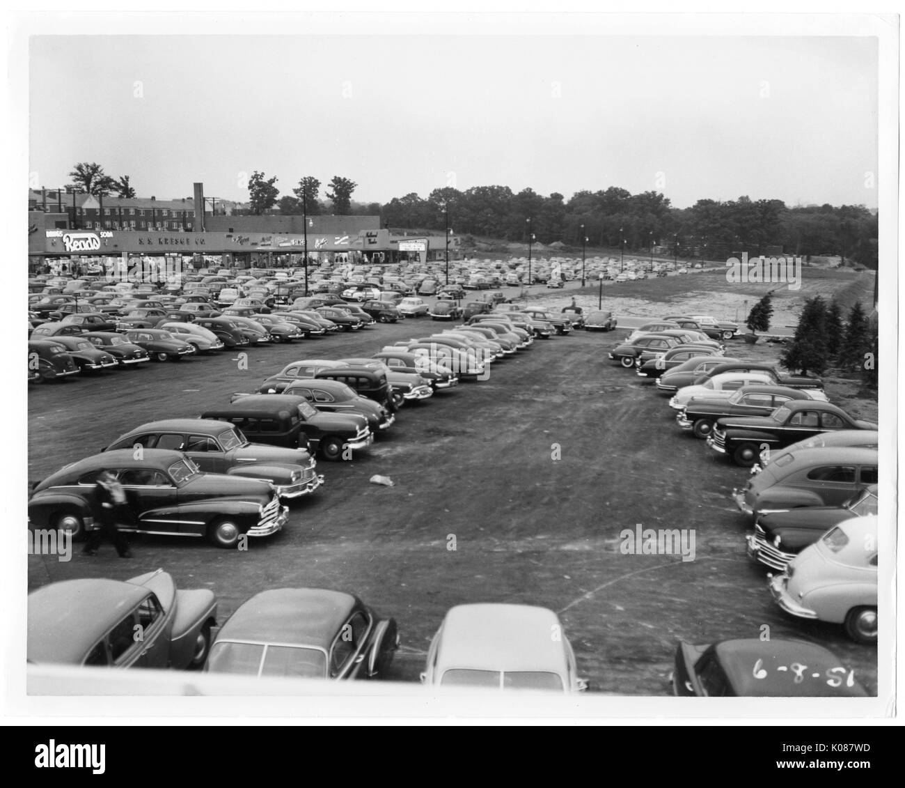 View of a packed parking lot that is under construction, in the background is part of the Northwood Shopping Center and behind the center are row homes to the left and trees to the right, one person is walking towards their car in the foreground, Baltimore, Maryland, 1951. Stock Photo