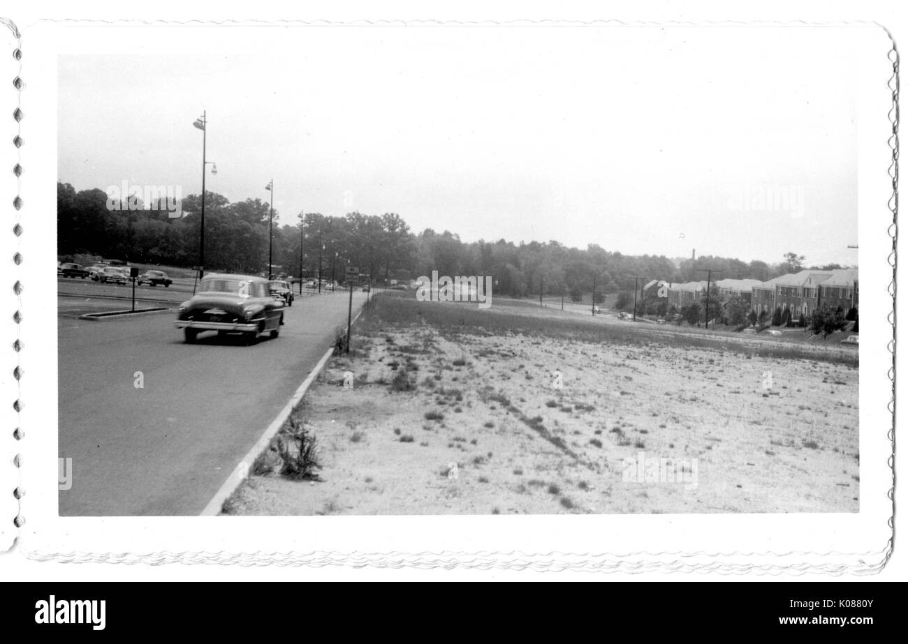 View of the large grass and dirt patch that is located between the Northwood Shopping Center and the row homes, one car on the shopping center side is driving towards the trees are in the background, Baltimore, Maryland, 1951. Stock Photo