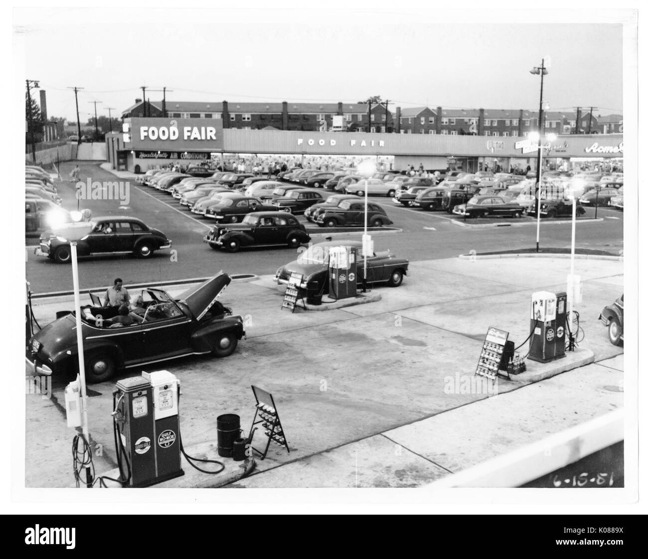 View of a gas station with two of the four pumps connected to cars, behind the gas station is a packed parking lot for the Food Fair store, behind the Food Fair store are row homes, Baltimore, Maryland, 1951. Stock Photo