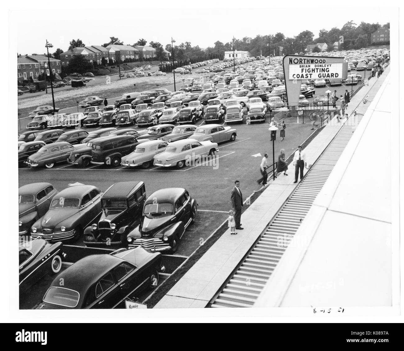 View of a packed parking lot from the roof of the Northwood Shopping Center, across the street from the center and parking lot are brick row homes and trees, people are heading to or from their cars, the center's sign is advertising 'FIGHTING COAST GUARD', Baltimore, Maryland, 1951. Stock Photo