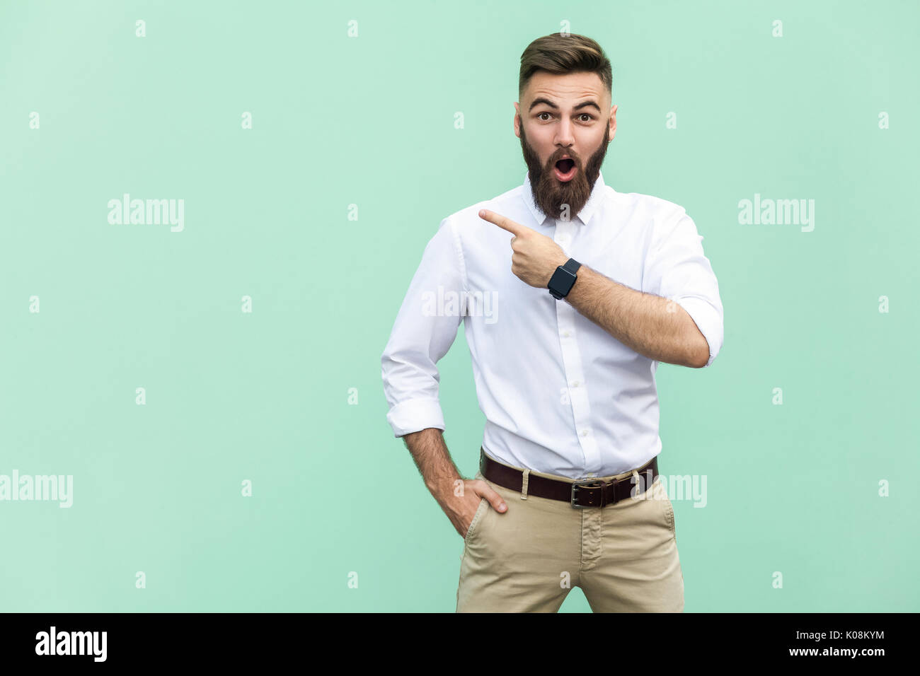 Wow! Handsome young adult man with beard in shoked. Pointing away while standing isolated on light green background Stock Photo