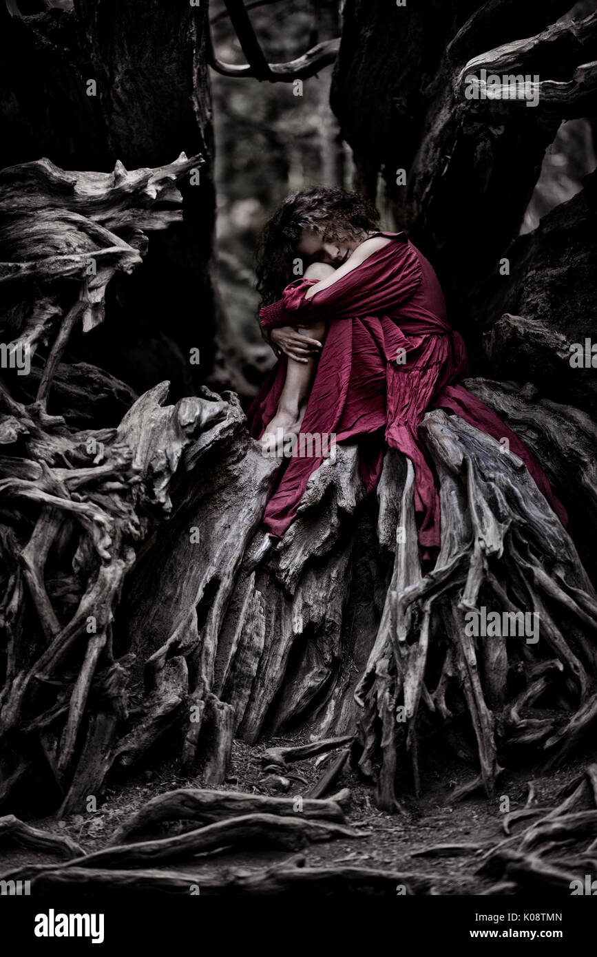 Beautiful young woman in a red dress curled up like a baby inside the root system of an old dead tree in a forest Stock Photo