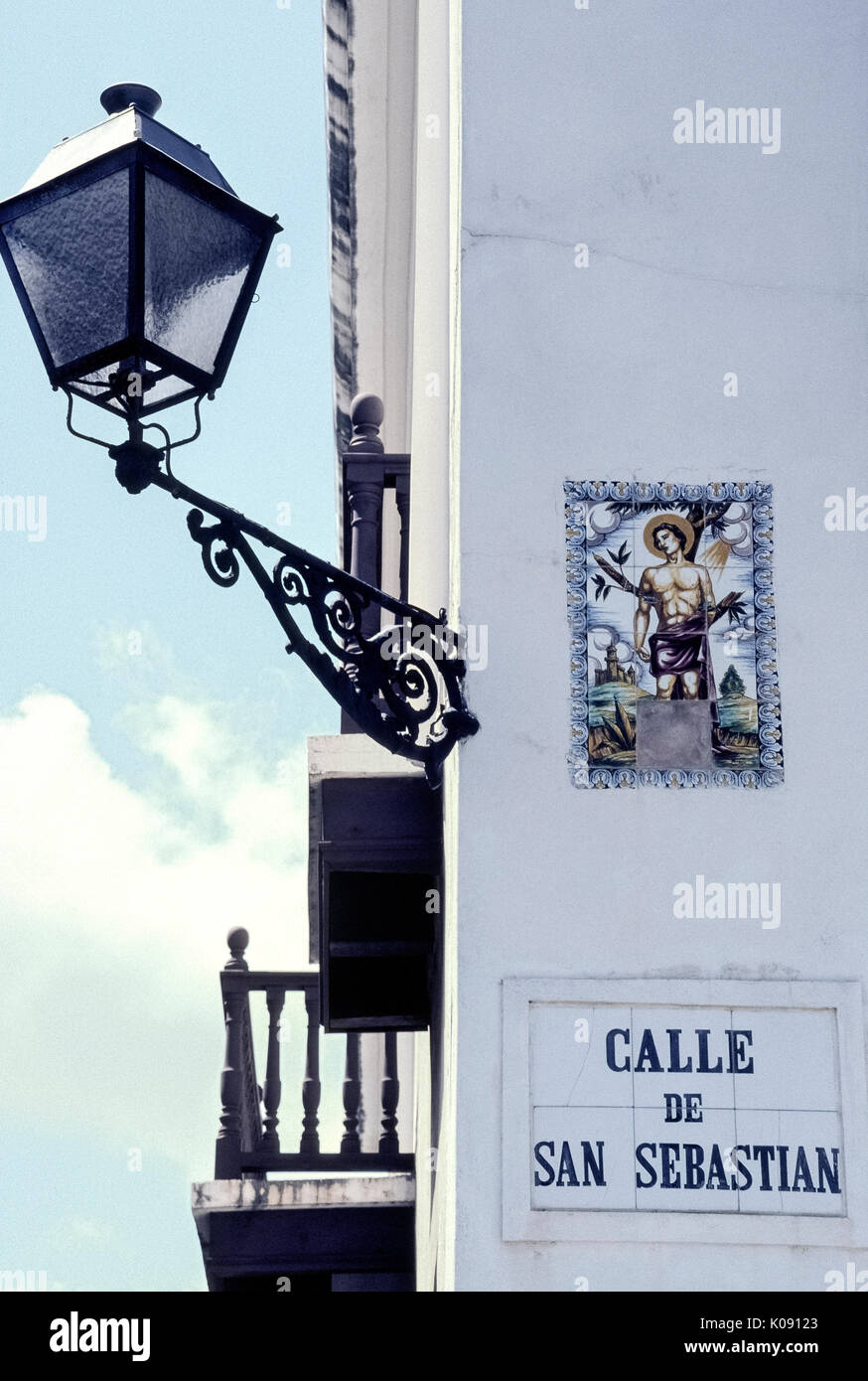 A religious artwork of ceramic tiles marks Calle de San Sebastian, a street named after early Christian martyr Saint Sebastian in historic Old San Juan in Puerto Rico (PR), an unincorporated territory of the United States in the Caribbean Sea. One of the tiles showing the saint's lower legs is missing. A vintage wrought iron street light illuminates the street name and illustrated sign at night. Stock Photo