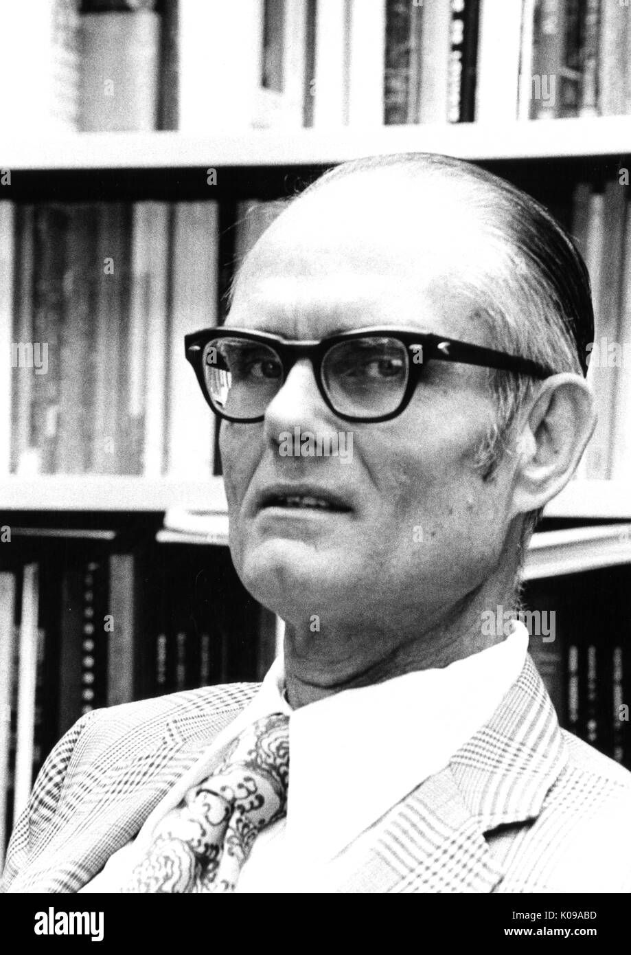 Head-shot of psychologist Julian Stanley, wearing a patterned suit and tie with a white shirt and dark thick glasses, seated in front of a bookshelf filled with books, with a serious facial expression. 1973. Stock Photo